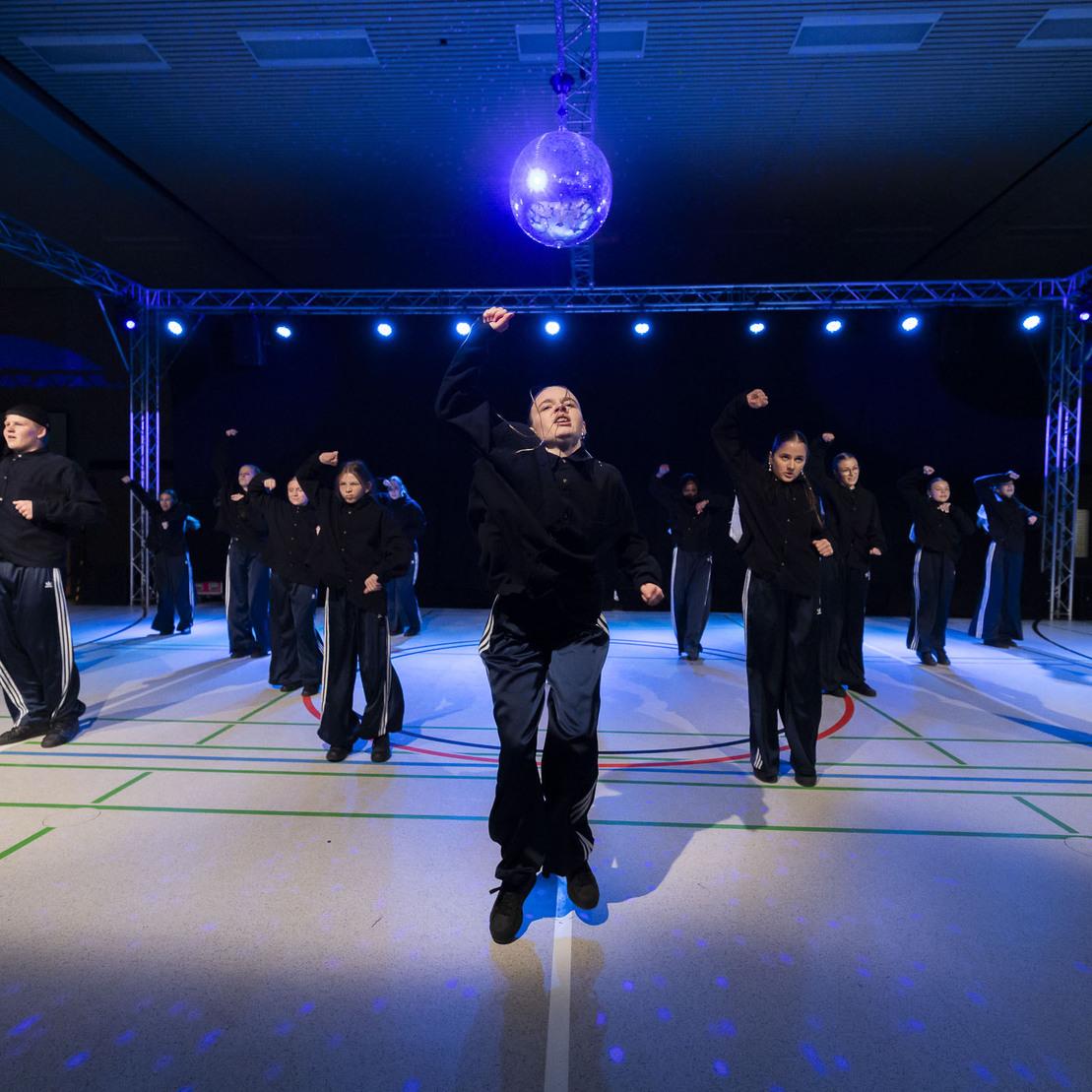 Tanzgruppe in schwarzen Outfits f&uuml;hrt synchronisierte Bewegungen unter einer Discokugel auf.