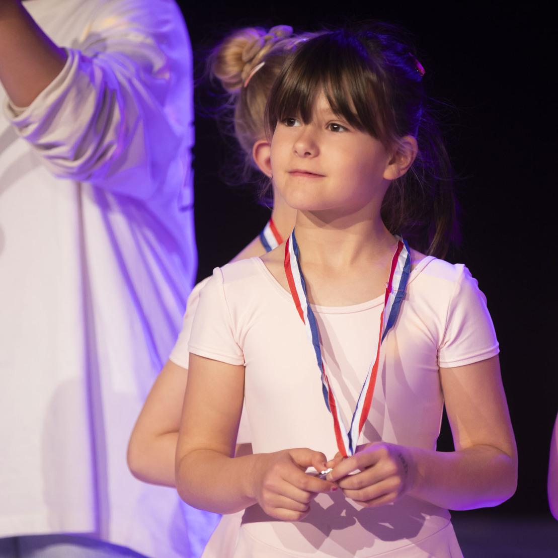 M&auml;dchen in rosa Tanzkleidung mit Medaille steht auf der B&uuml;hne, blickt stolz und h&auml;lt eine zweite Medaille in der Hand.