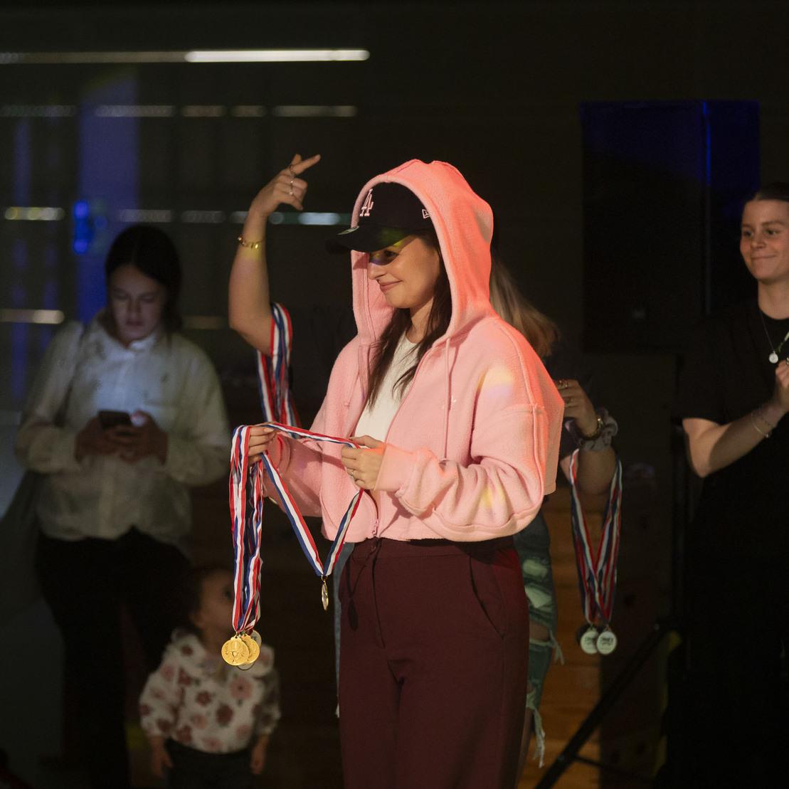 Junge Frau in pinkem Hoodie erh&auml;lt Medaillen bei einer Preisverleihung, umgeben von applaudierenden Personen.