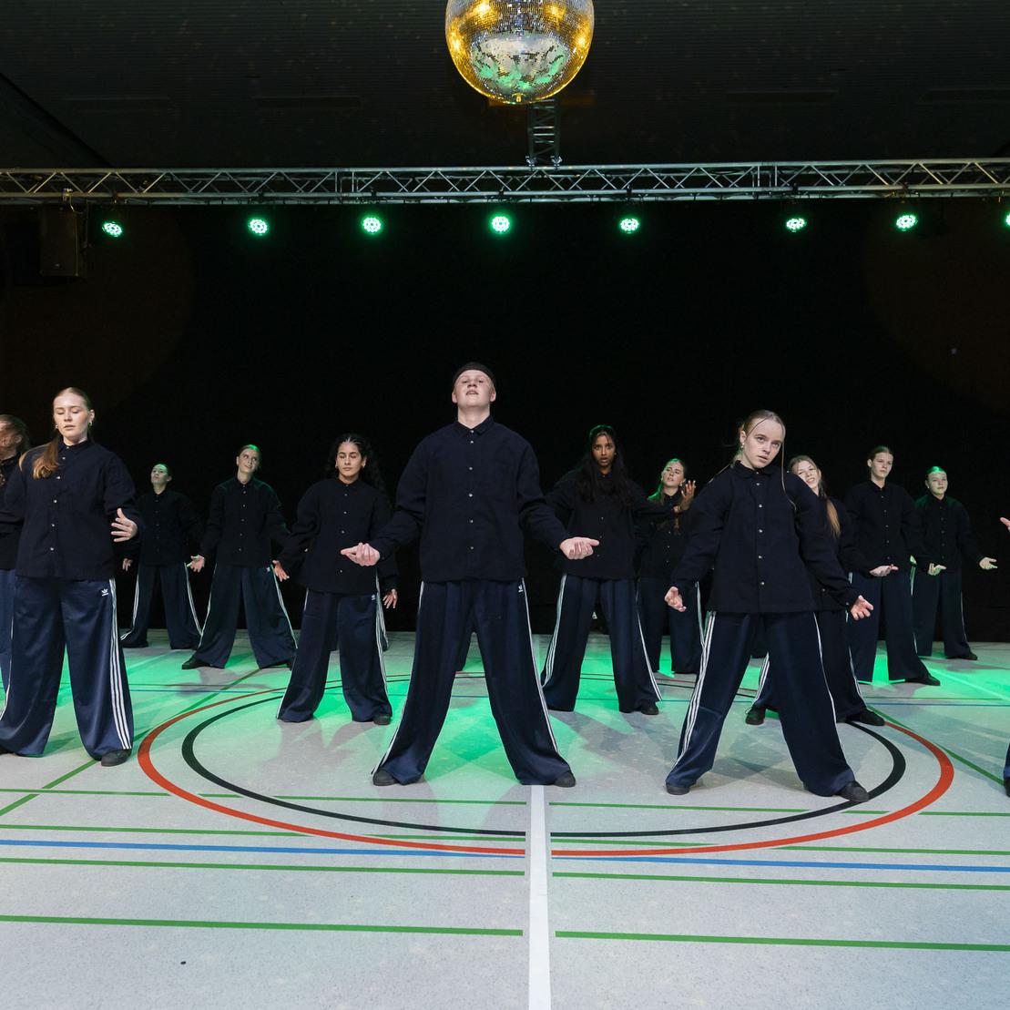 Tanzgruppe in schwarzen Outfits f&uuml;hrt synchron eine Choreografie auf einer beleuchteten B&uuml;hne mit Discokugel vor.