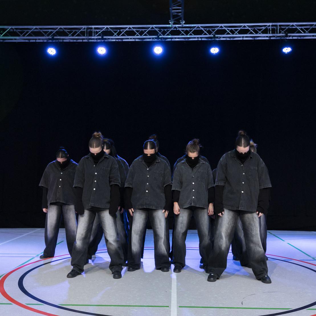 Sechs T&auml;nzer in schwarzen Outfits stehen auf einem Sportboden, beleuchtet von blauen Scheinwerfern.