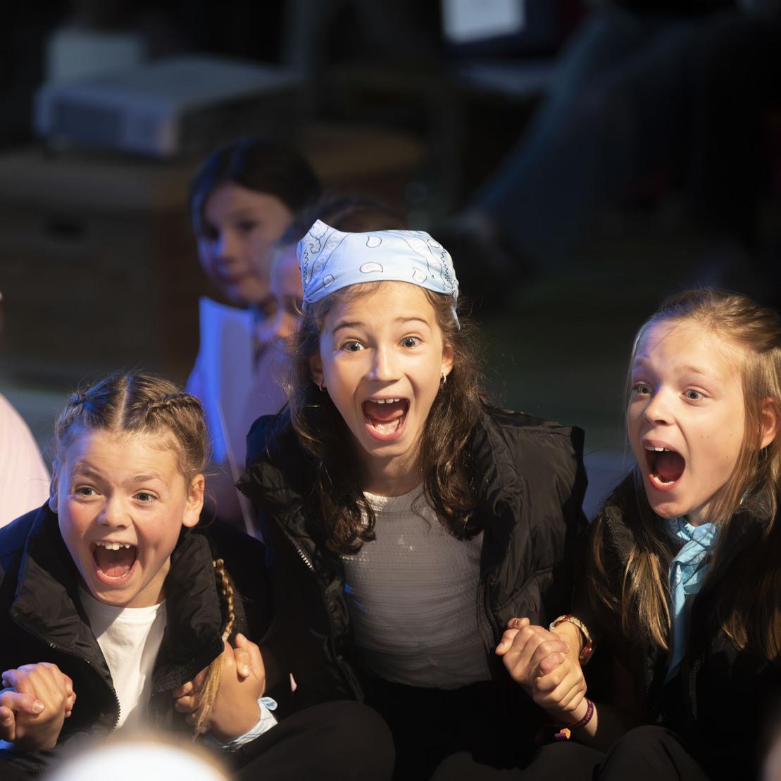 Gruppe von aufgeregten Kindern sitzt zusammen und jubelt begeistert, einige mit weit ge&ouml;ffnetem Mund und fr&ouml;hlichen Gesichtsausdr&uuml;cken.