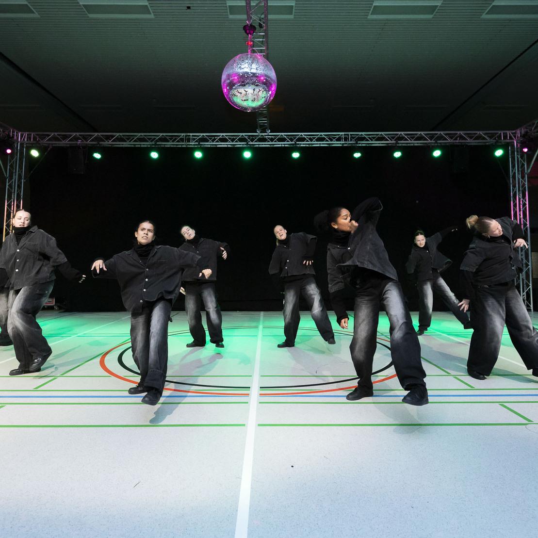 Tanzgruppe in schwarzen Outfits f&uuml;hrt eine Choreografie auf einer Sportfl&auml;che mit Diskokugel und Beleuchtung vor.