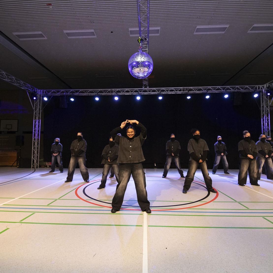Tanzgruppe mit einheitlicher Kleidung f&uuml;hrt Performance unter einer Discokugel auf einer Sporthalle auf.