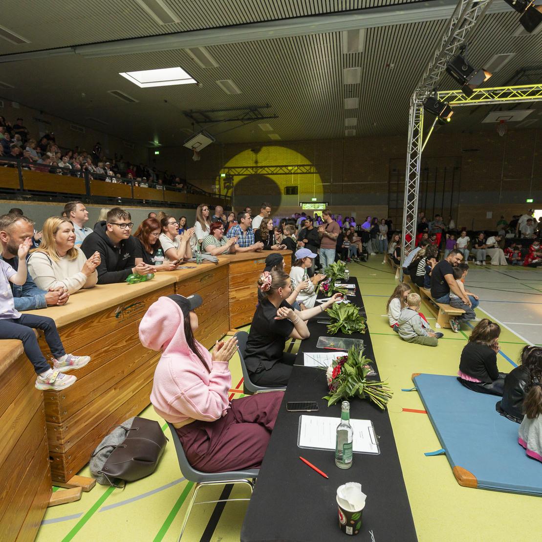 Zuschauer applaudieren in einer Sporthalle, w&auml;hrend eine Jury am Tisch sitzt und eine Veranstaltung beobachtet.
