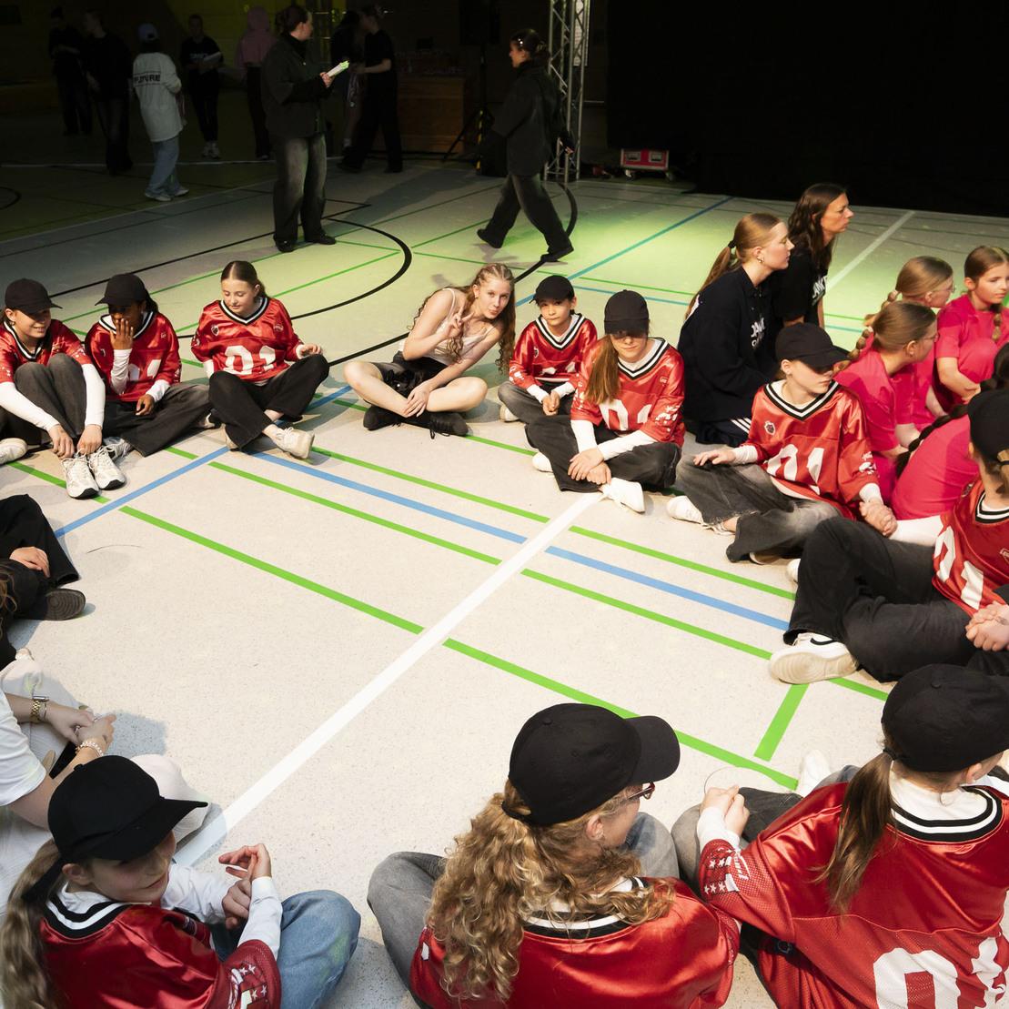 Gruppenszenen von Kindern in roten Trikots und Caps, die im Halbkreis auf dem Boden sitzen und sich unterhalten.
