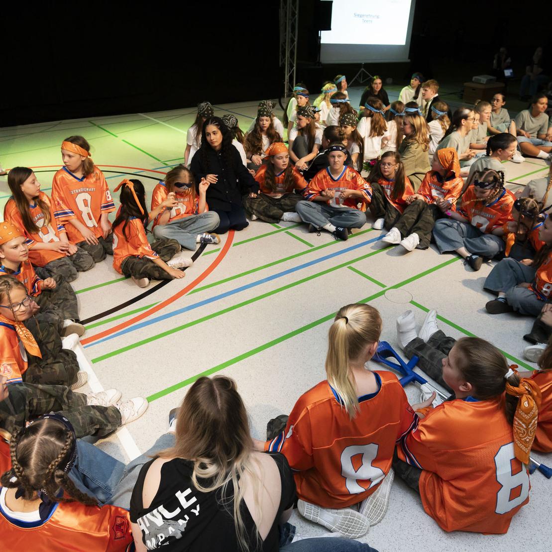 Gruppen von Kindern in orangefarbenen Trikots sitzen in einem Hallenraum auf dem Boden, w&auml;hrend eine Person spricht.