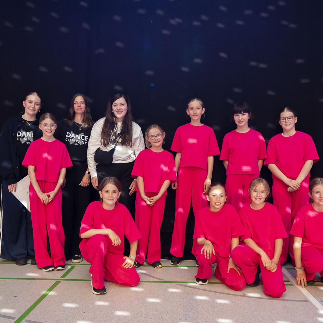 Gruppe von T&auml;nzern in roten Outfits posiert vor schwarzem Hintergrund mit Lichtreflexionen.