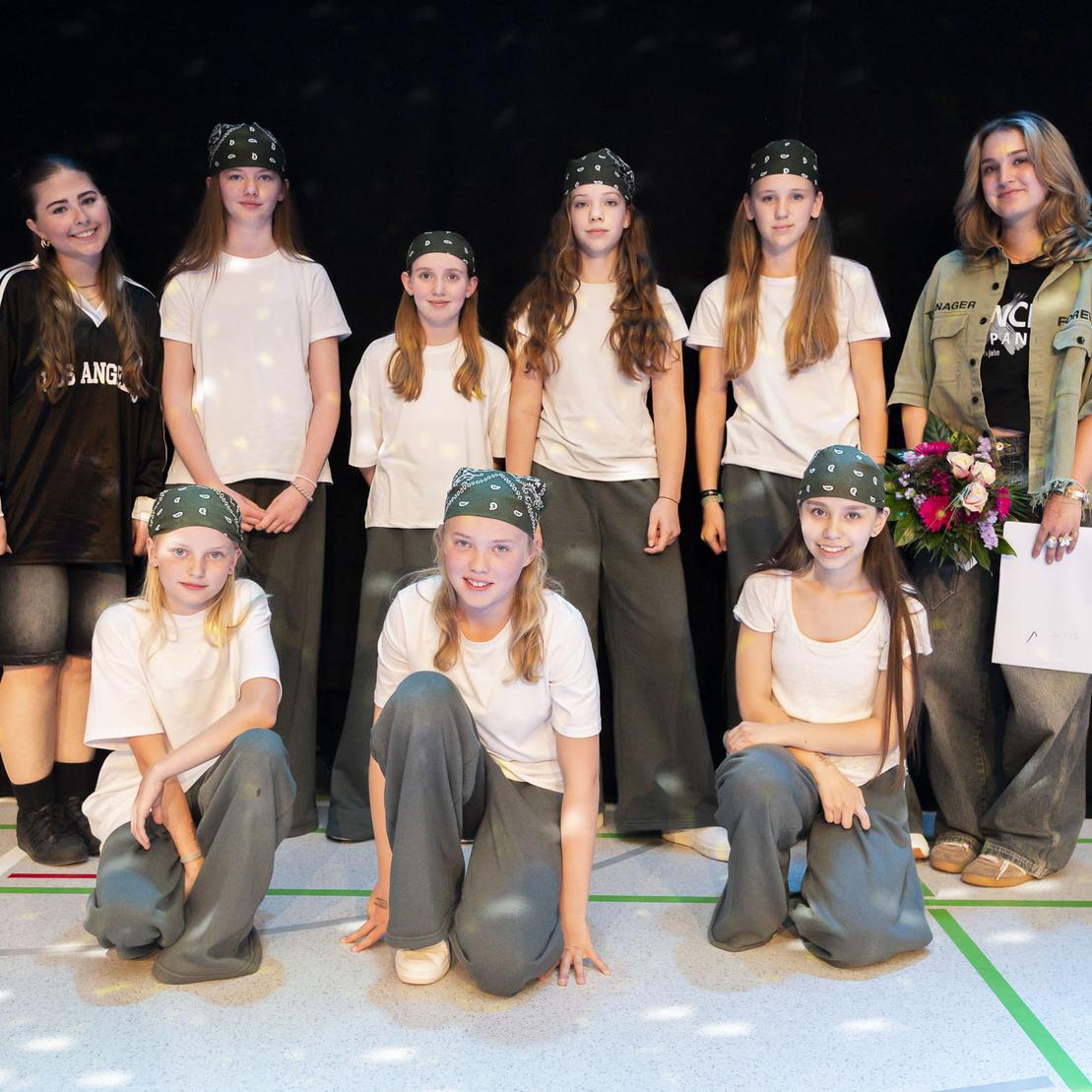 Gruppe von sieben T&auml;nzerinnen in &auml;hnlicher Kleidung, die auf einer B&uuml;hne posieren, umrahmt von einer Choreografin.