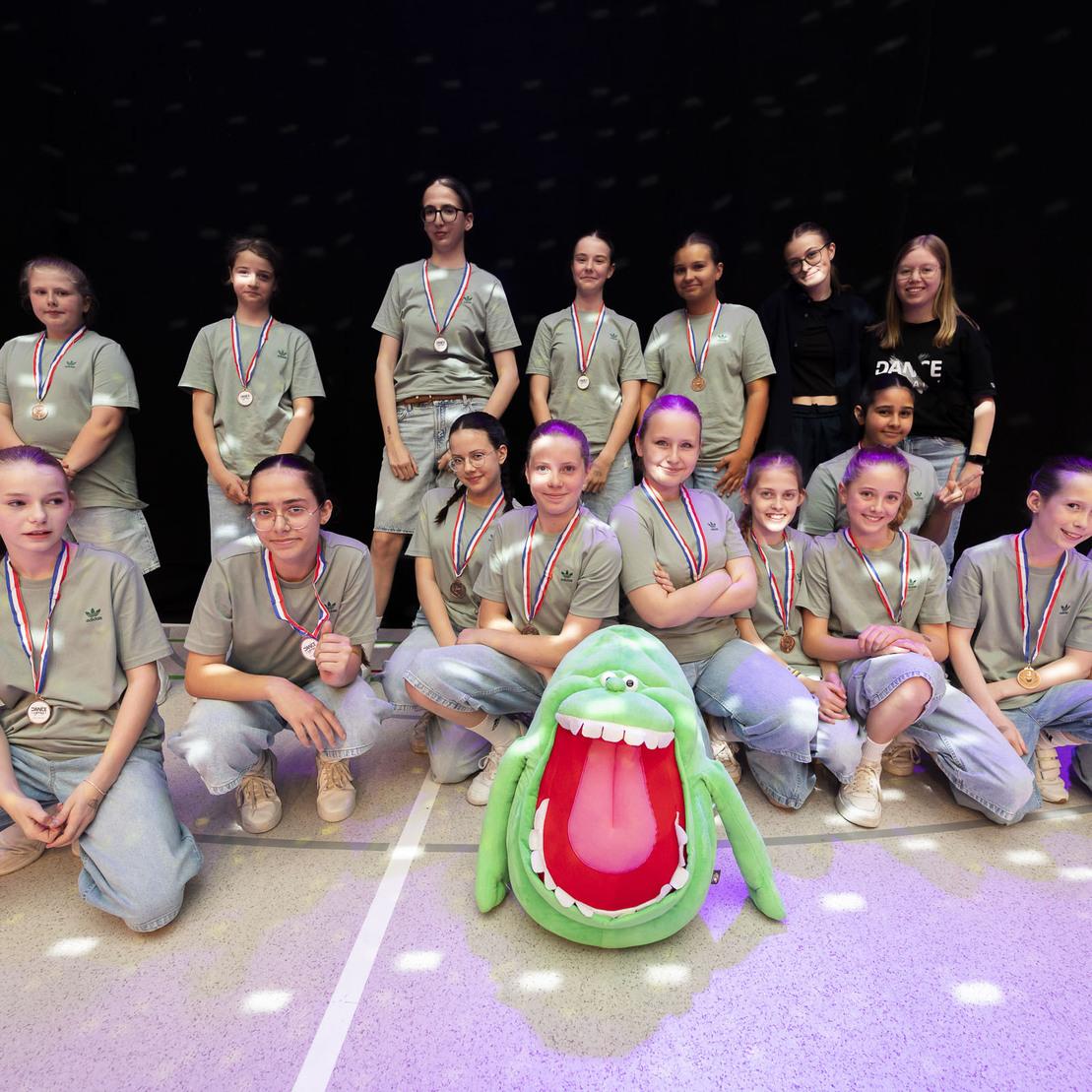 Gruppe von zw&ouml;lf M&auml;dchen in grauen T-Shirts mit Medaillen, posierend vor einem gro&szlig;en, gr&uuml;nen Pl&uuml;schmonster.