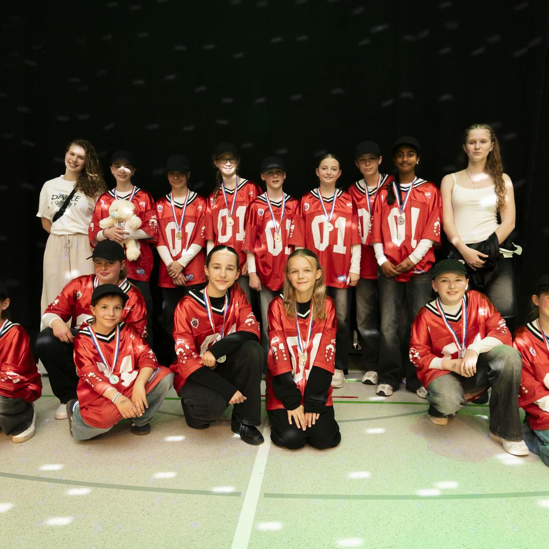 Gruppennfoto von 15 Kindern in roten Jacken mit Medaillen, umgeben von buntem Licht, auf einem Sportboden.