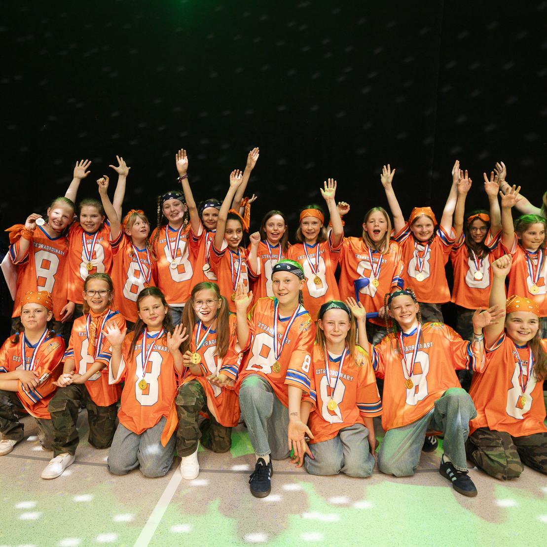Gruppe von Kindern in orangefarbenen Trikots mit Medaillen, die jubelnd auf einer B&uuml;hne stehen.