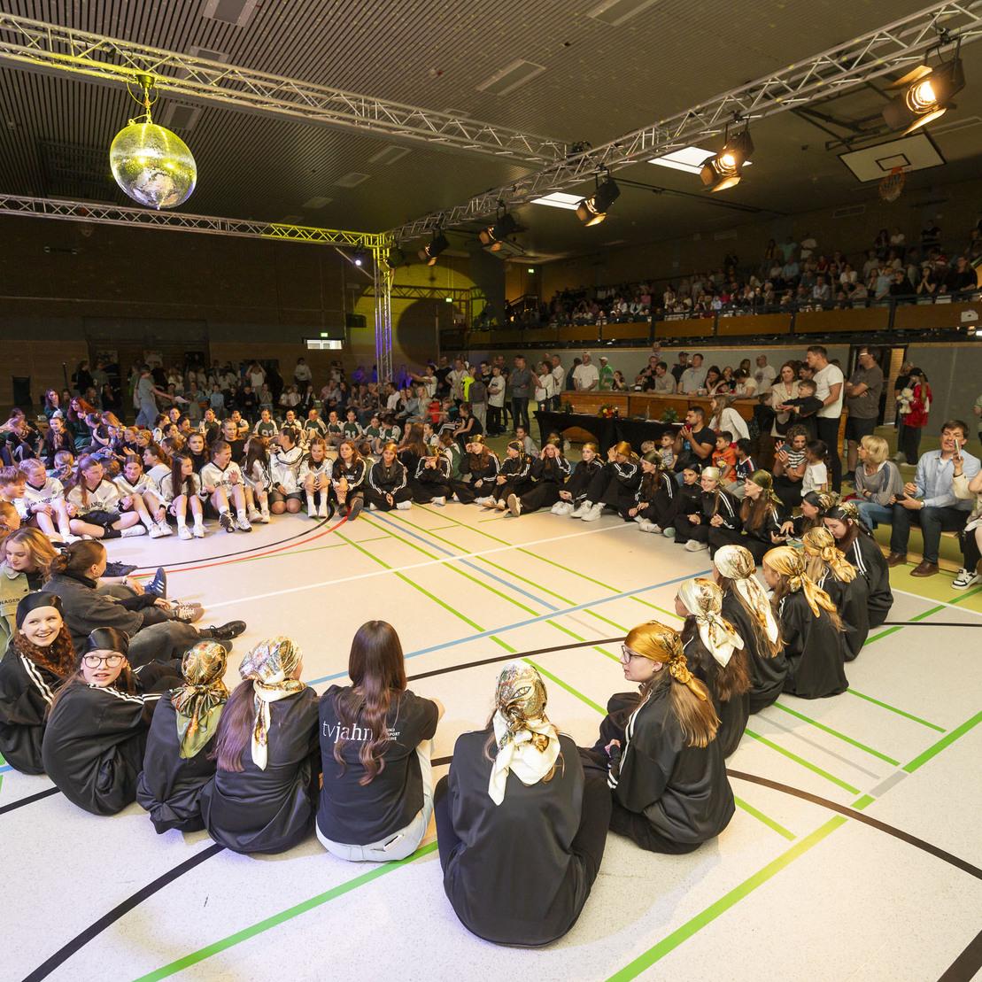 Gruppenvorf&uuml;hrung in einer Sporthalle mit Kindern und Jugendlichen, die im Kreis sitzen, umringt von Zuschauern.