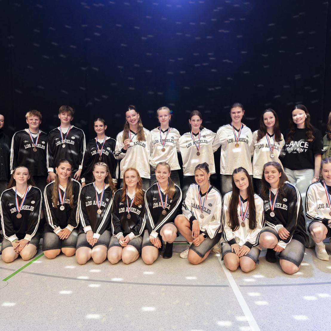 Gruppe von T&auml;nzern in Sportkleidung mit Medaillen in einer festlichen Atmosph&auml;re vor dunklem Hintergrund.