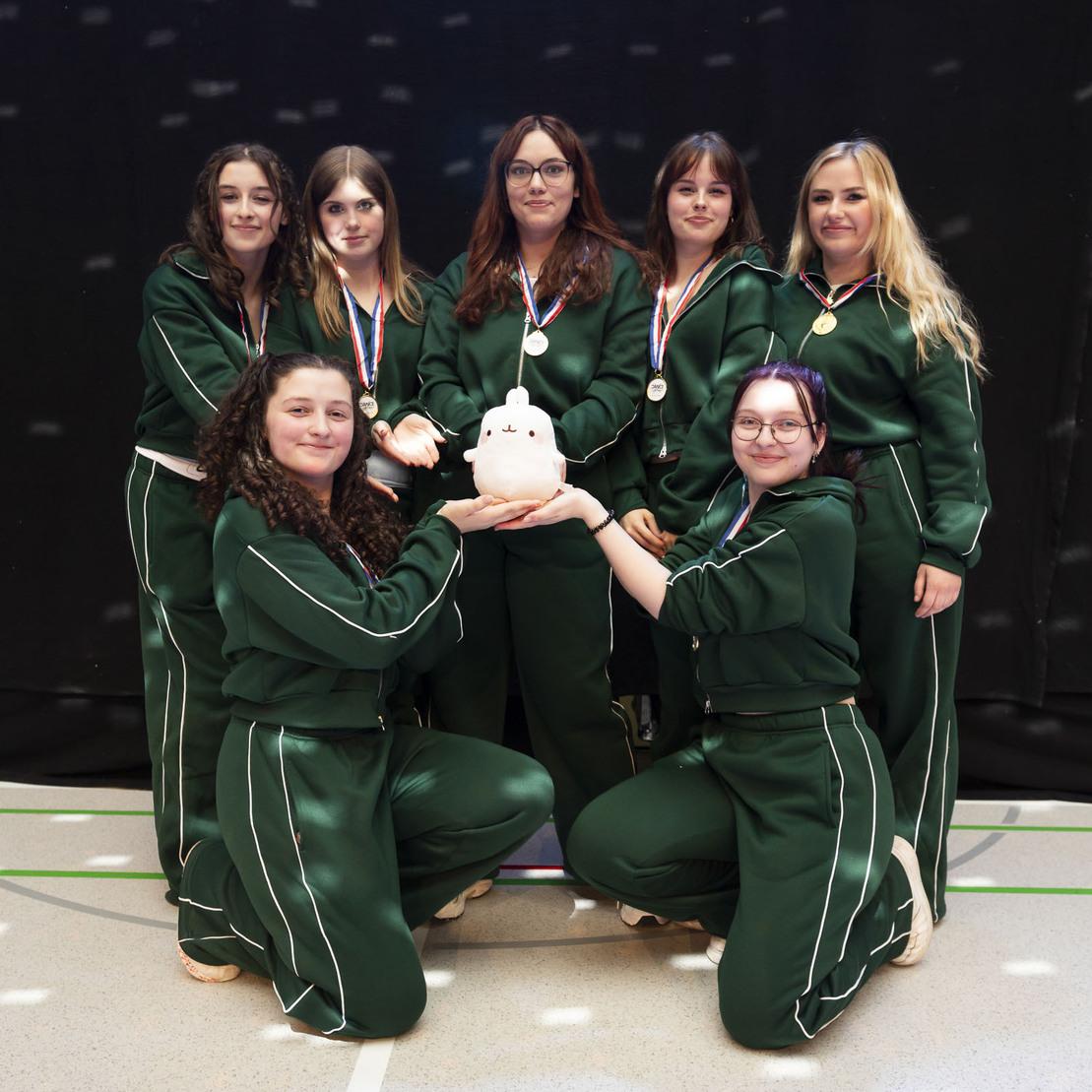 Gruppenfoto von sieben Frauen in gr&uuml;nen Trainingsanz&uuml;gen, die gemeinsam eine Sparschweinfigur halten.