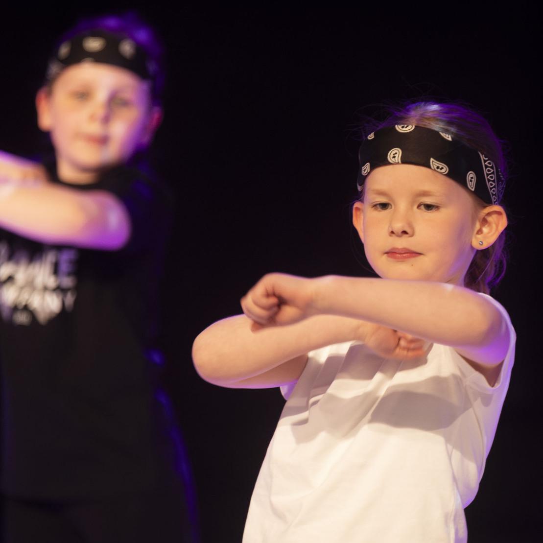Zwei Kinder mit Bandanas f&uuml;hren eine Tanzbewegung auf einer B&uuml;hne aus, mit verschwommenem Hintergrund.