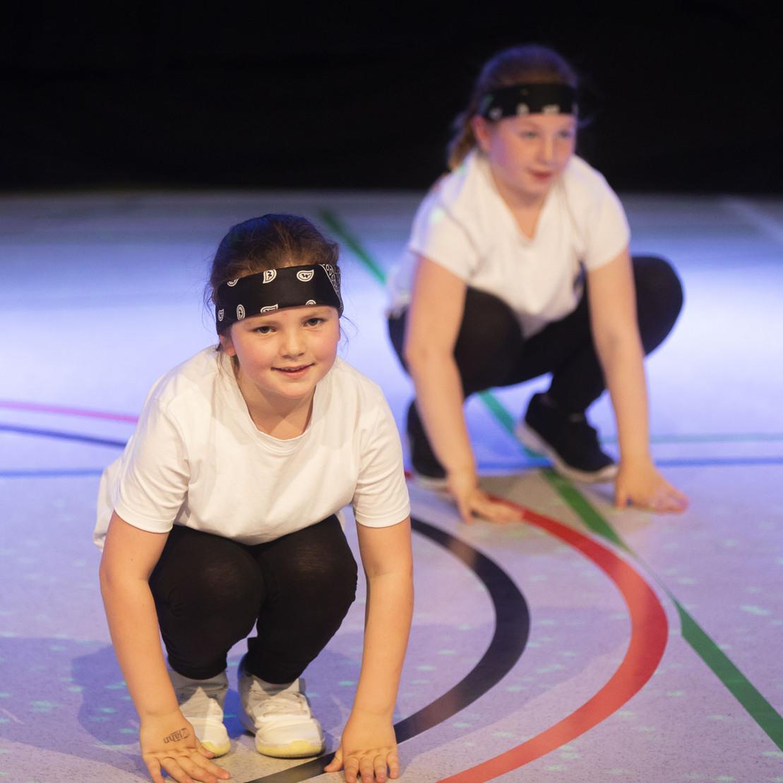 Zwei Kinder in sportlichen Outfits mit Stirnb&auml;ndern, in der Hocke auf einer bunten Gymnastikfl&auml;che.