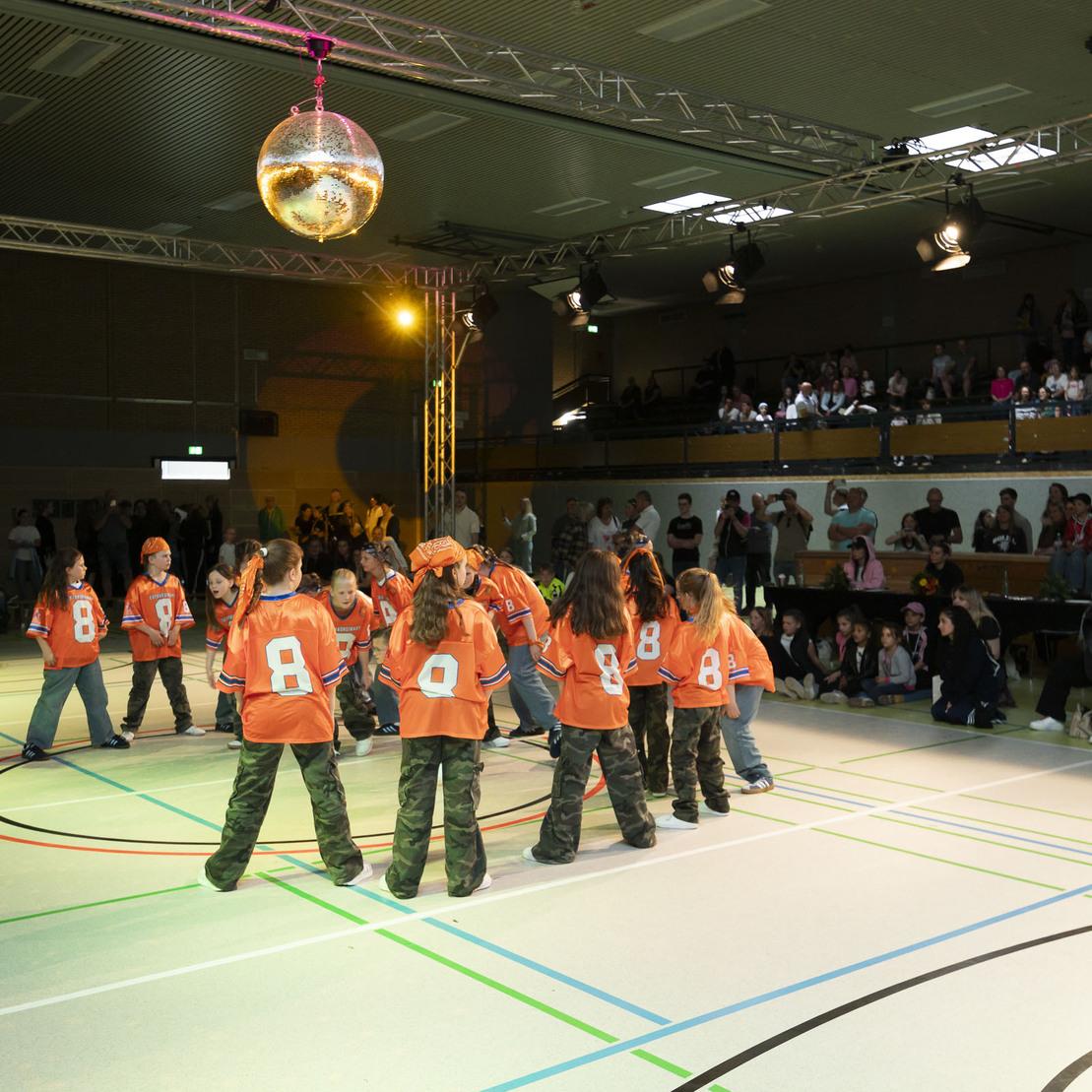 Jugendliche in orangefarbenen Shirts tanzen auf einer Sportfl&auml;che unter einer Discokugel, Publikum schaut zu.