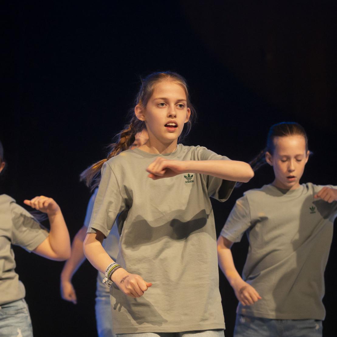 Tanzgruppe von M&auml;dchen in grauen T-Shirts f&uuml;hrt eine choreografierte Tanzroutine auf einer B&uuml;hne vor.
