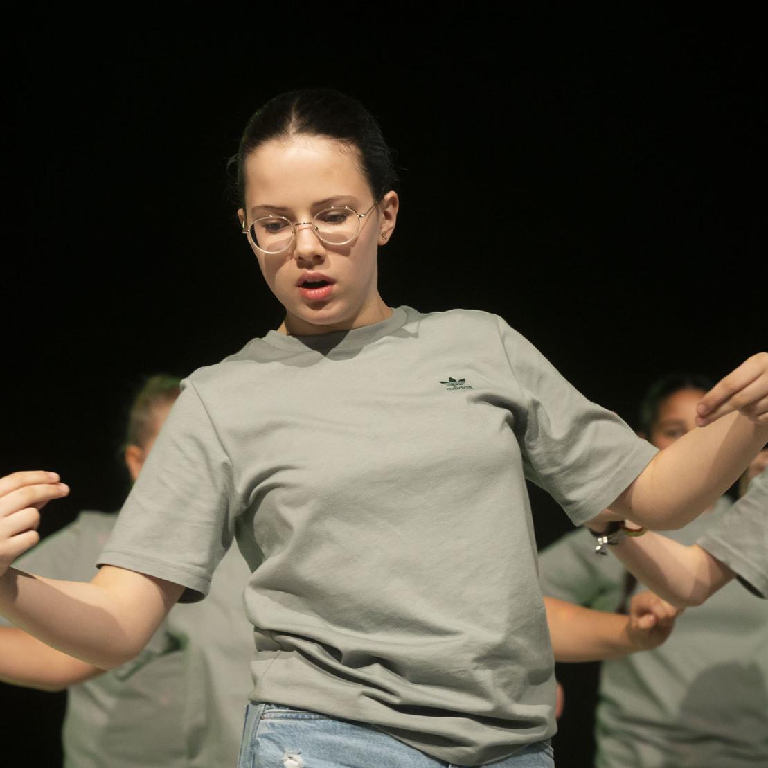 M&auml;dchen tanzt auf der B&uuml;hne, tr&auml;gt ein graues T-Shirt und Jeans, mit konzentriertem Gesichtsausdruck.
