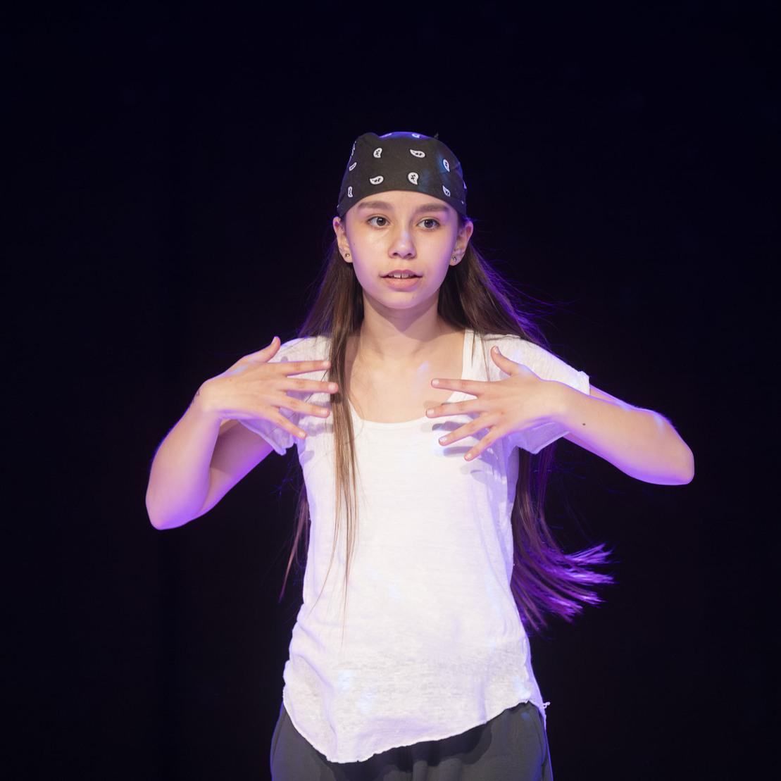 M&auml;dchen mit schwarzem Bandana und wei&szlig;em Shirt, steht im Spotlight und gestikuliert w&auml;hrend einer Tanzperformance.