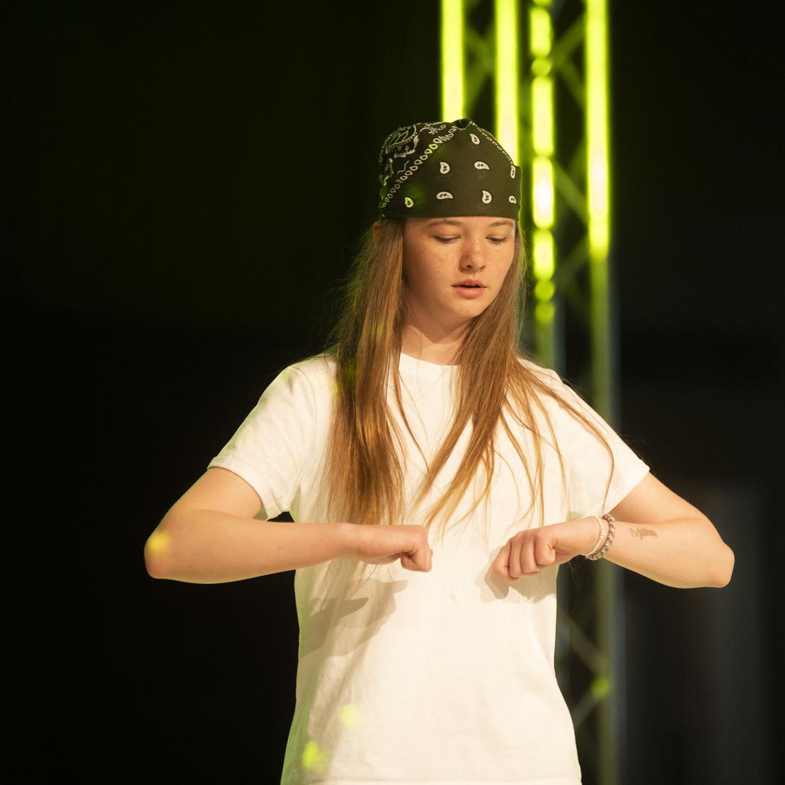 T&auml;nzerin in wei&szlig;em T-Shirt und Bandana pr&auml;sentiert eine Performance auf einer B&uuml;hne mit bunten Lichtern.