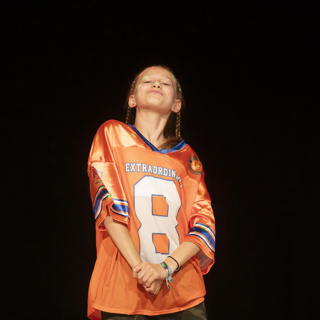 Ein Kind in einem orangefarbenen Trikot mit der Nummer 8 posiert selbstbewusst auf einer dunklen B&uuml;hne.