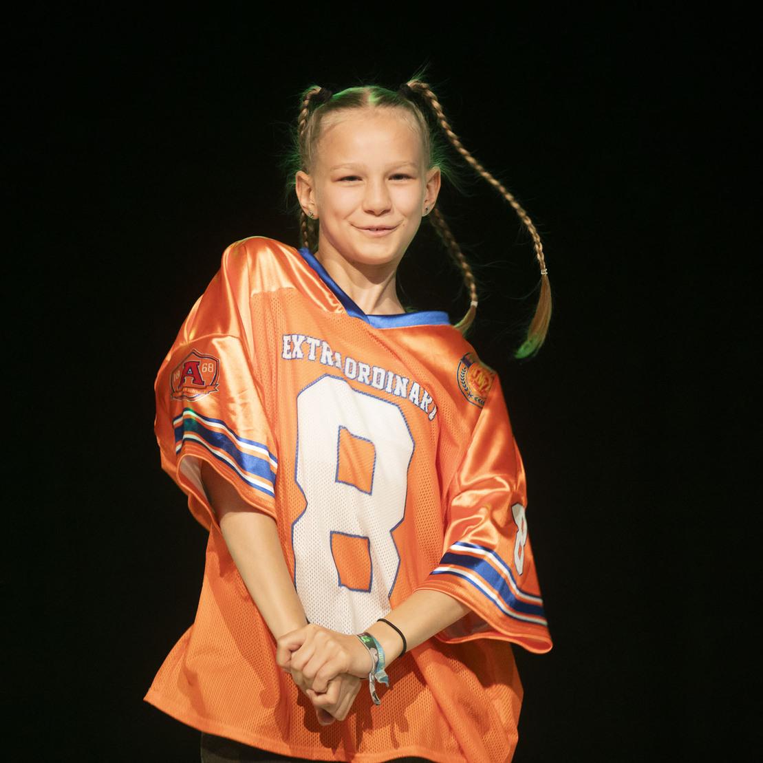 M&auml;dchen mit Geflochtenen Z&ouml;pfen tr&auml;gt ein orangefarbenes Trikot mit der Nummer 8 und l&auml;chelt auf einer B&uuml;hne.