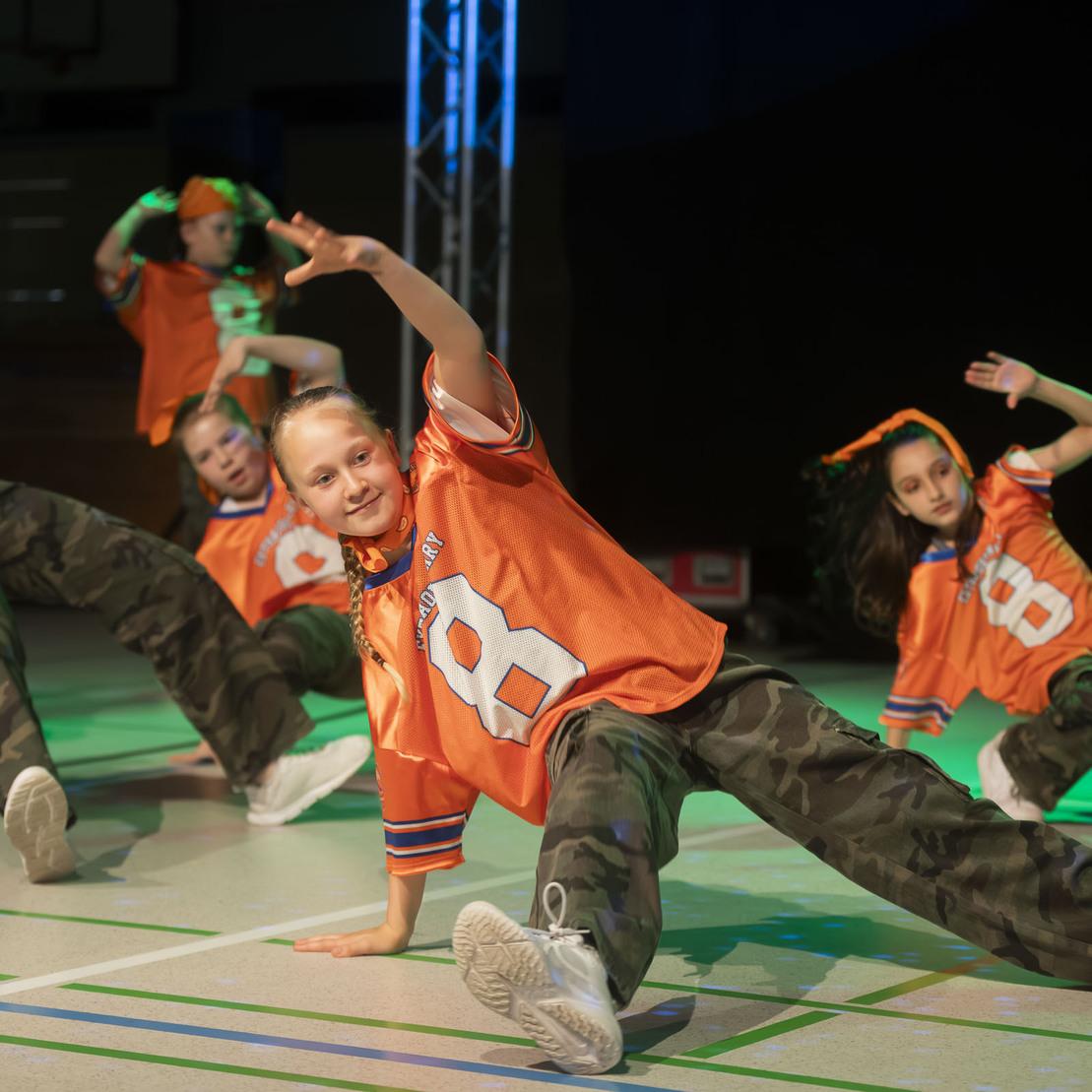 Tanzgruppe in orangefarbenen Trikots f&uuml;hrt eine Pose auf einer Sportfl&auml;che mit bunten Lichtern aus.