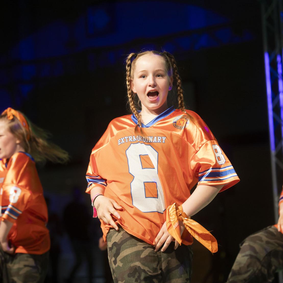 Drei T&auml;nzerinnen in orangefarbenen Trikots mit der Nummer 8 und Camouflage-Hosen bei einer Performance auf einer B&uuml;hne.