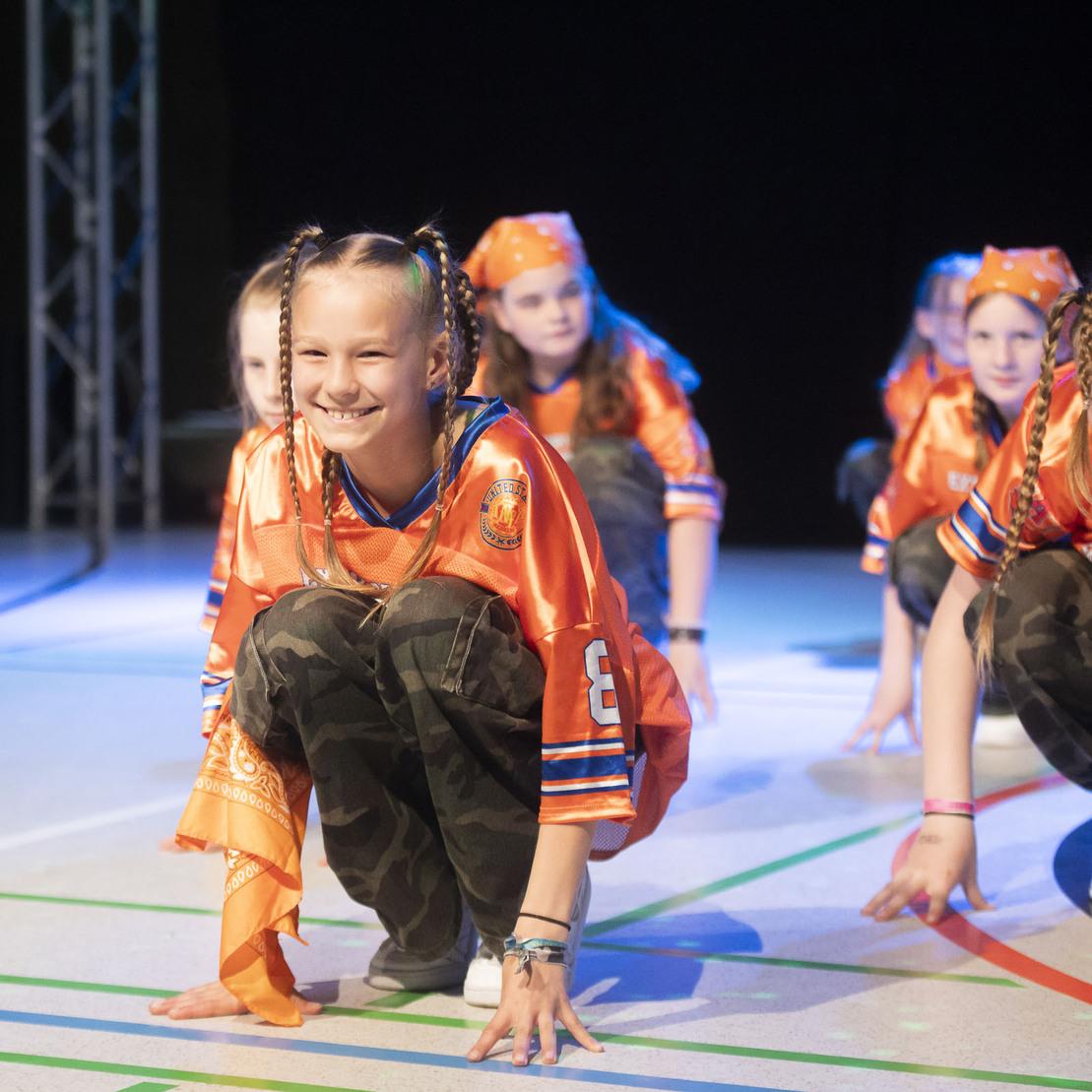Tanzende Kinder in orangefarbener Kleidung f&uuml;hren eine Choreografie auf einer B&uuml;hne mit buntem Licht aus.