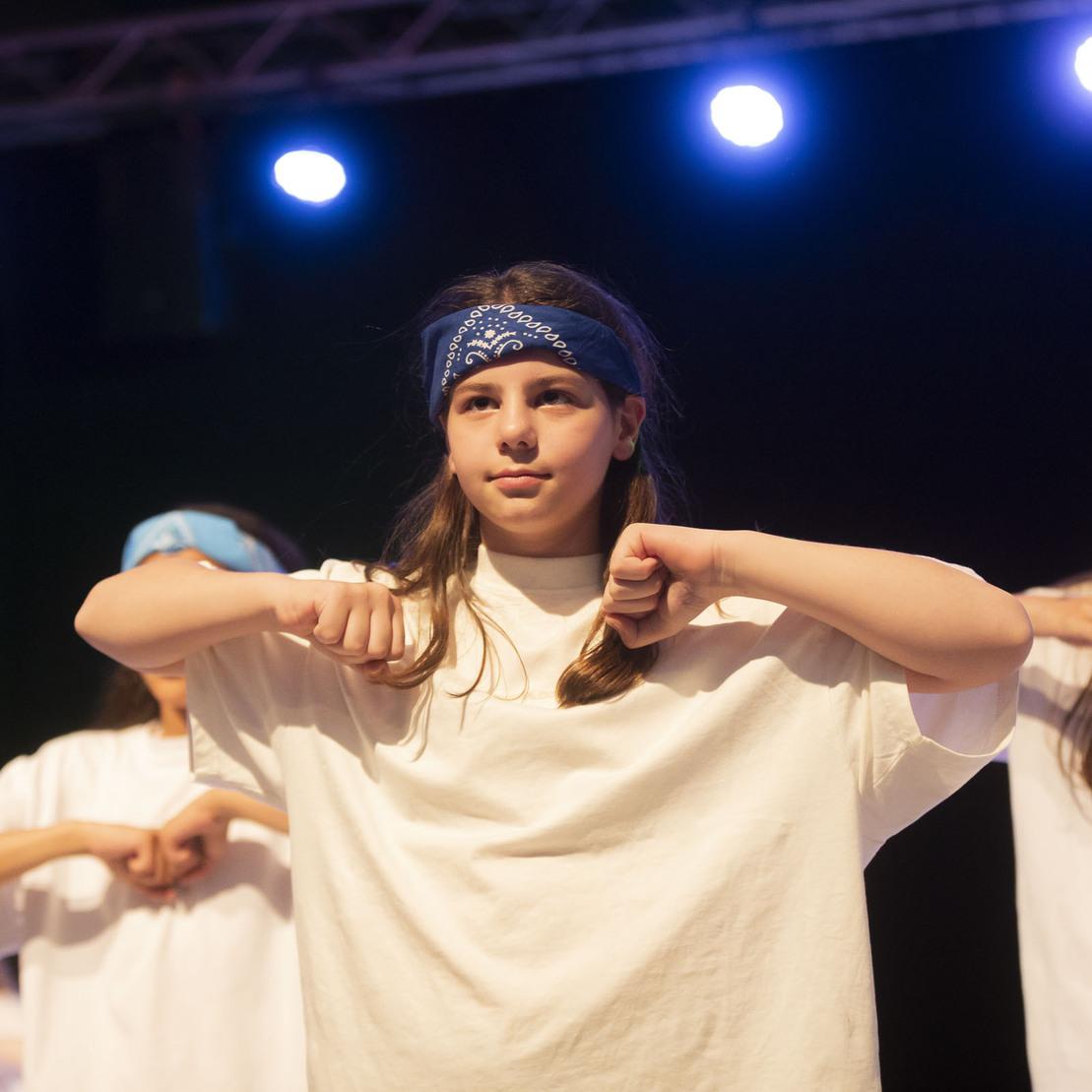 Tanzende Kinder in wei&szlig;en T-Shirts und blauen Haarb&auml;ndern f&uuml;hren eine synchronisierte Choreografie auf einer B&uuml;hne vor.