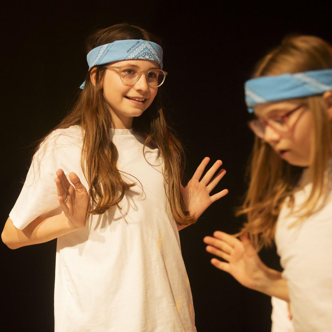 Zwei M&auml;dchen in wei&szlig;en T-Shirts und blauen Bandanas interagieren auf einer B&uuml;hne.