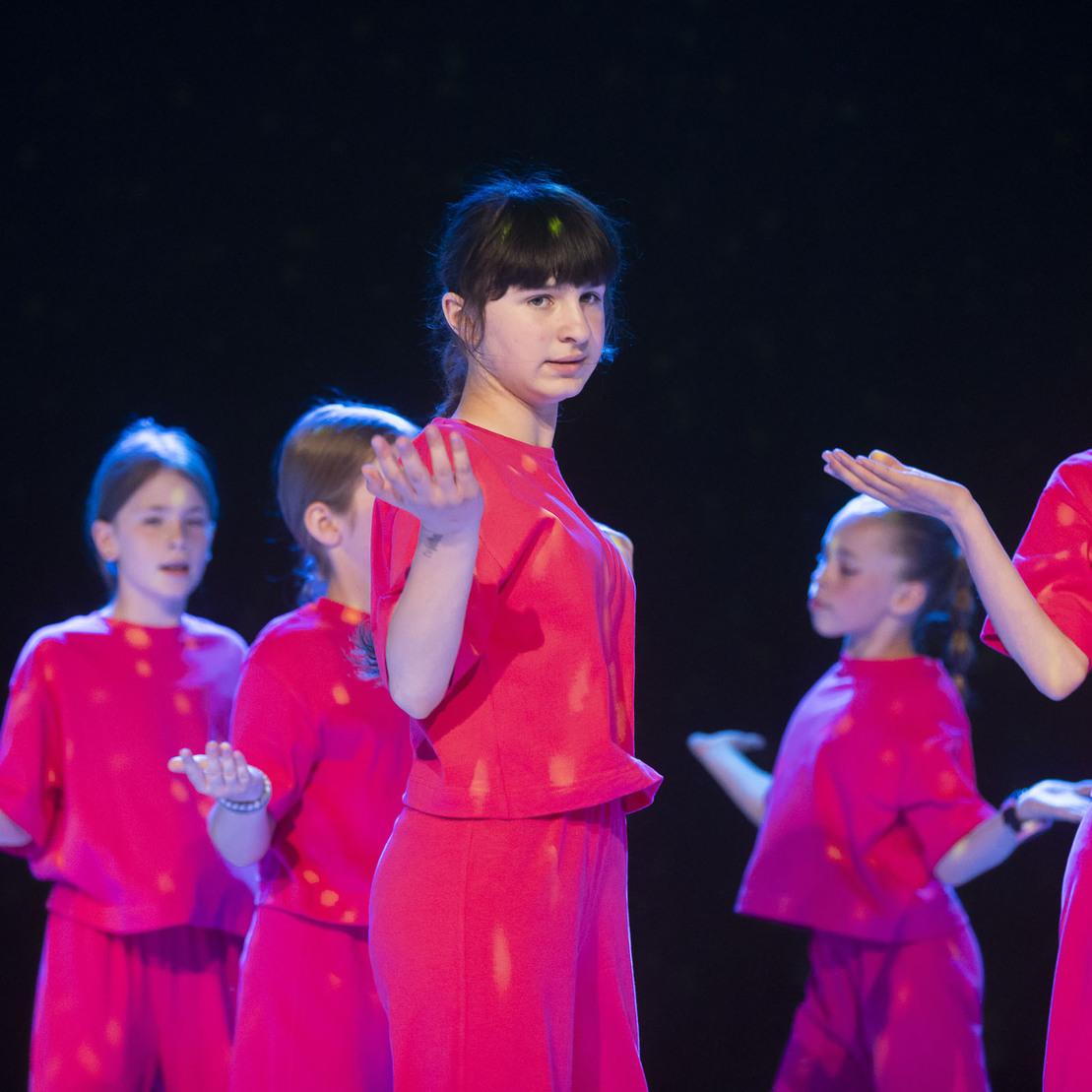 Sechs T&auml;nzerinnen in roten Outfits f&uuml;hren eine Choreografie auf einer dunklen B&uuml;hne auf.