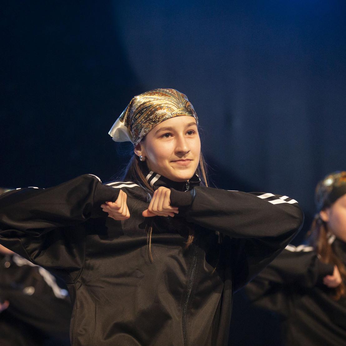 T&auml;nzerin in schwarzer Jacke mit Kopftuch, die auf einer B&uuml;hne eine Gruppenperformance zeigt.