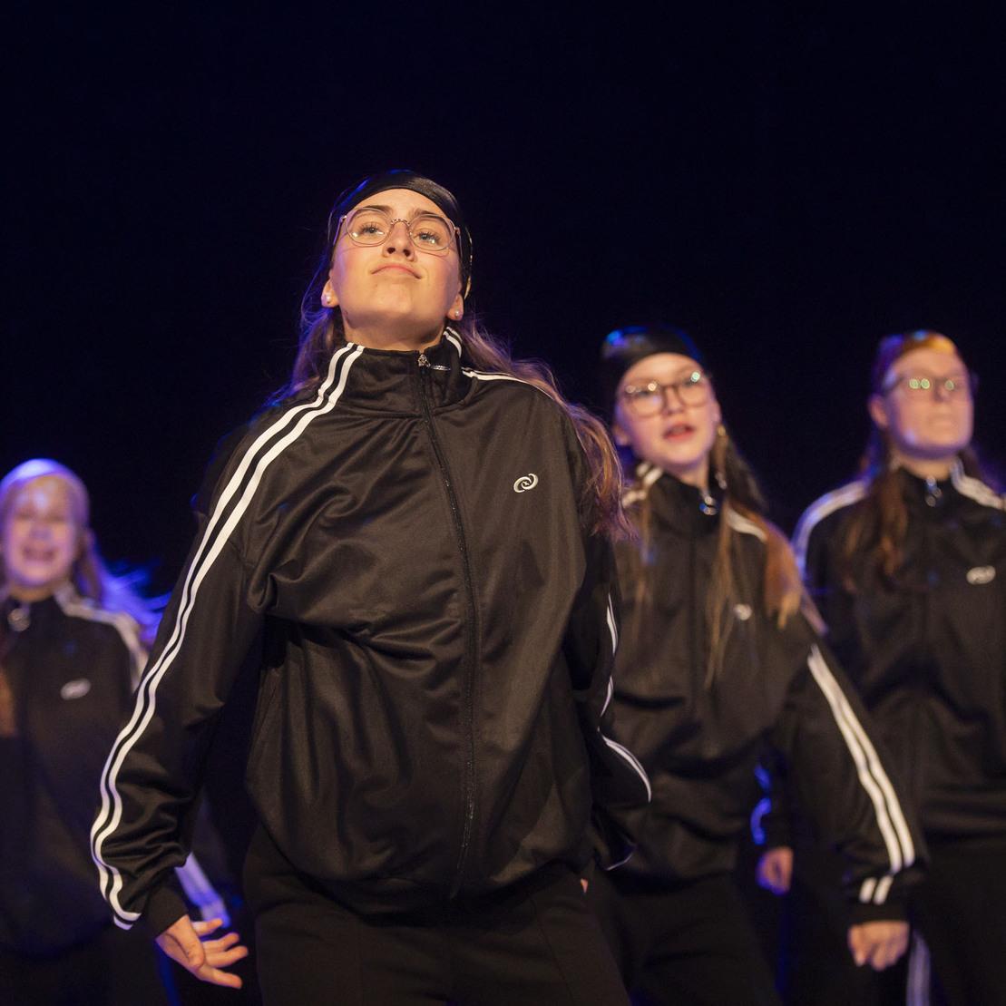 T&auml;nzerinnen in schwarzen Trainingsanz&uuml;gen f&uuml;hren eine Choreografie auf einer dunklen B&uuml;hne auf.