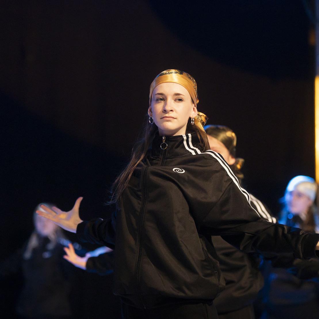 T&auml;nzerin in schwarzer Sportbekleidung mit goldener Kopfbedeckung, steht im Fokus auf einer B&uuml;hne mit Licht-Effekten.