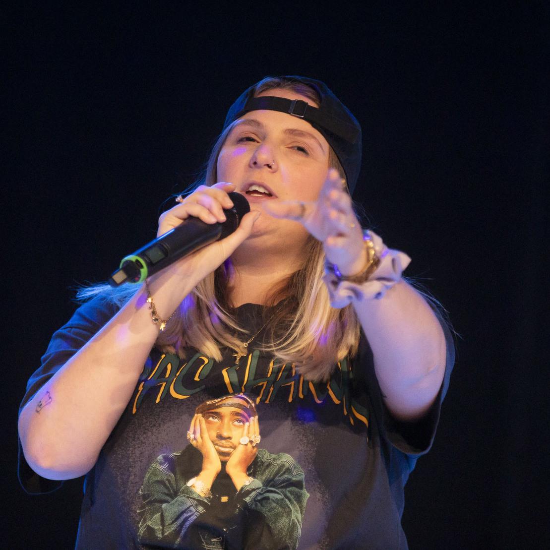 S&auml;ngerin mit Baseballcap h&auml;lt ein Mikrofon und performt auf einer dunklen B&uuml;hne, T-Shirt mit Bild von Tupac.