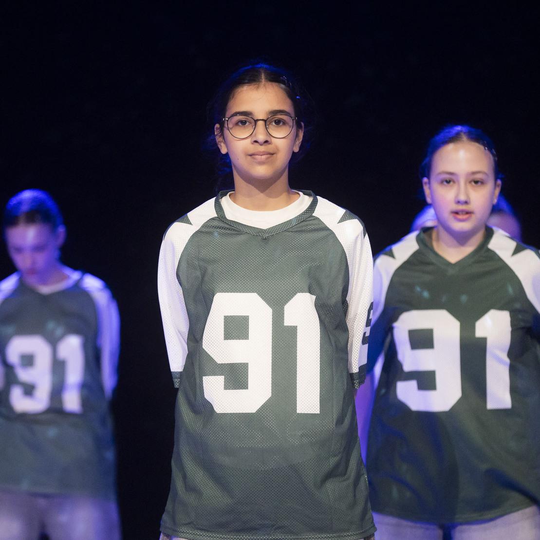 Gruppendarstellung von M&auml;dchen in sportlichen Trikots mit der Nummer 91 auf einer Theaterb&uuml;hne.