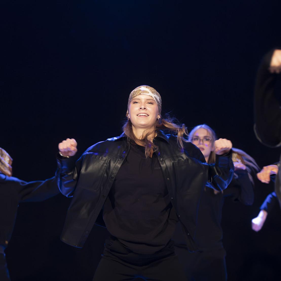 Tanzgruppe mit mehreren T&auml;nzerinnen in Schwarz, die synchron eine kraftvolle Choreografie auf einer B&uuml;hne auff&uuml;hren.