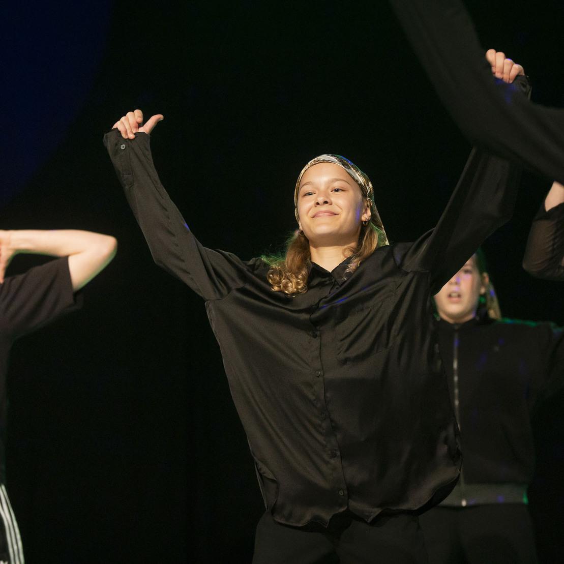 Tanzende Gruppe von M&auml;dchen in schwarzen Outfits, die fr&ouml;hlich auf einer B&uuml;hne performen.