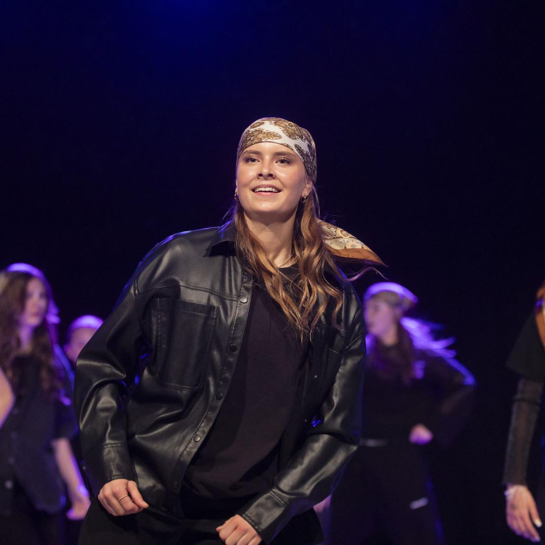 T&auml;nzerin l&auml;chelt w&auml;hrend einer Performance, tr&auml;gt eine schwarze Jacke und ein Kopftuch, mit weiteren T&auml;nzern im Hintergrund.
