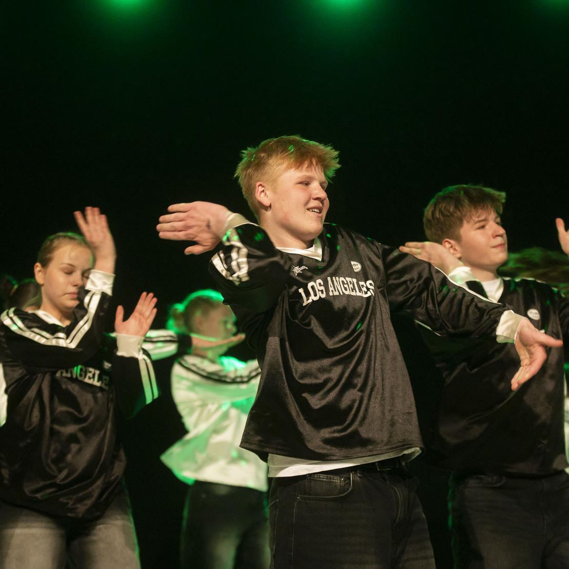 Tanzgruppe in sportlicher Kleidung f&uuml;hrt synchron eine Choreografie auf einer B&uuml;hne mit gr&uuml;ner Beleuchtung vor.