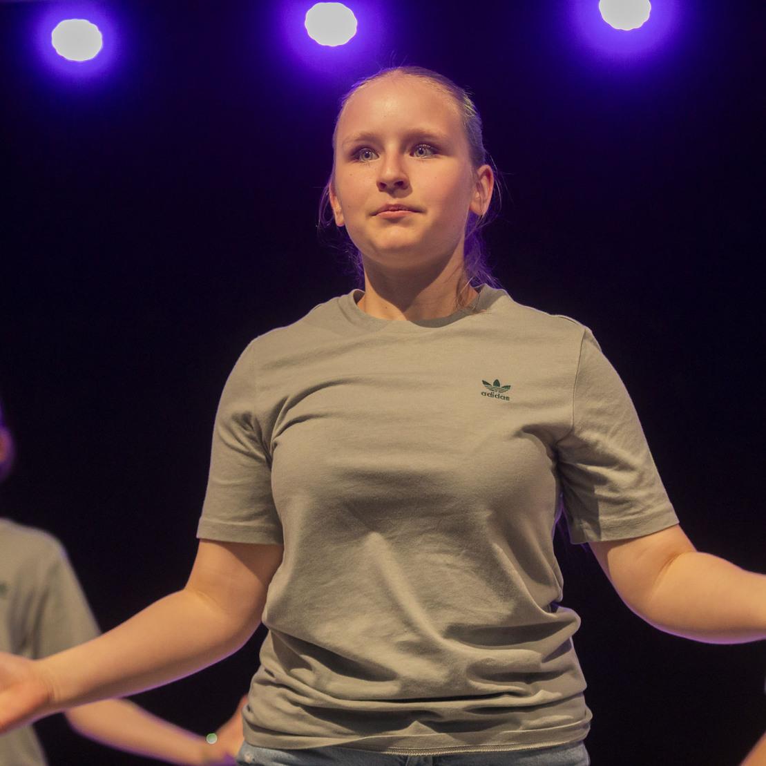 Junge T&auml;nzerin auf der B&uuml;hne, umgeben von zwei weiteren T&auml;nzerinnen, unter lila Beleuchtung in grauen T-Shirts.