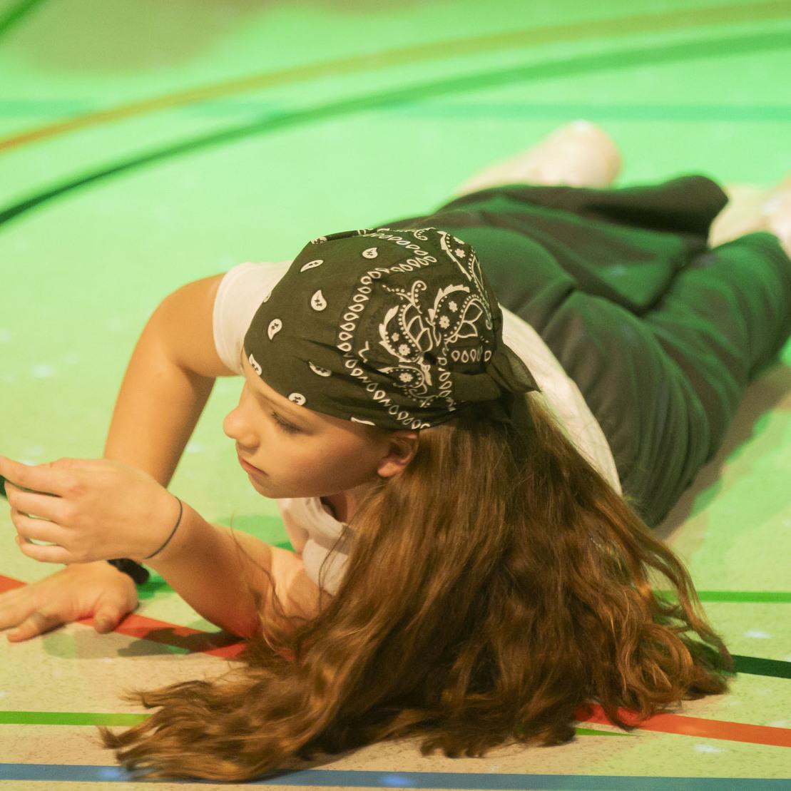 M&auml;dchen mit Bandana liegt auf dem Boden einer Sporthalle und gestikuliert auf einer bunten Linienfl&auml;che.
