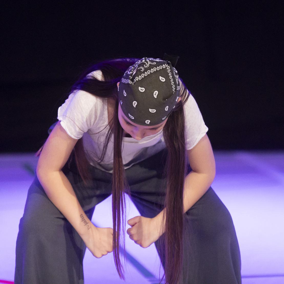 T&auml;nzerin mit Bandana beugt sich auf einer B&uuml;hne, umgeben von sanftem Licht in Violett- und Blaut&ouml;nen.