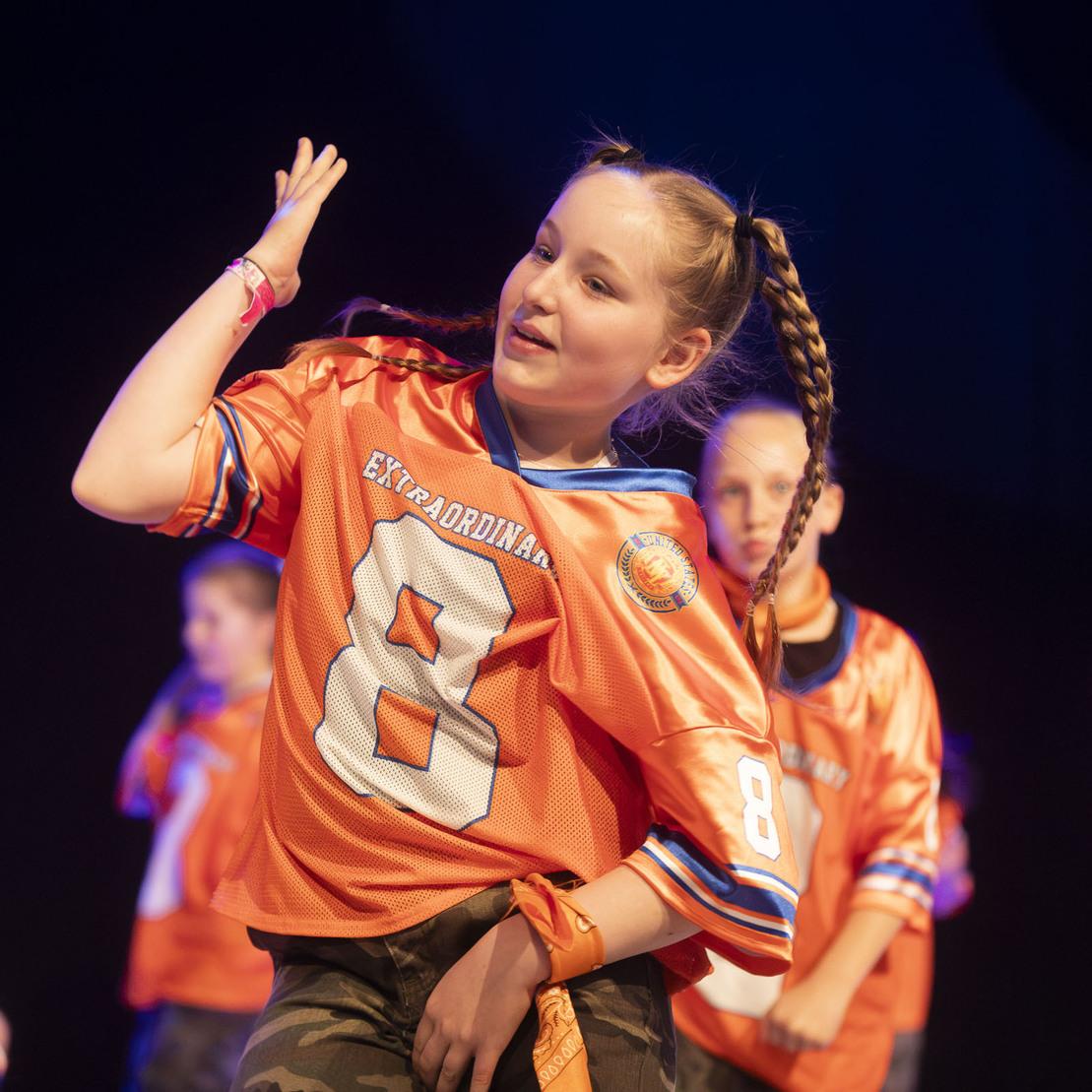 Tanzende Kinder in orangefarbenen Trikots auf der B&uuml;hne, eines hebt die Hand zu einem Tanzmove.