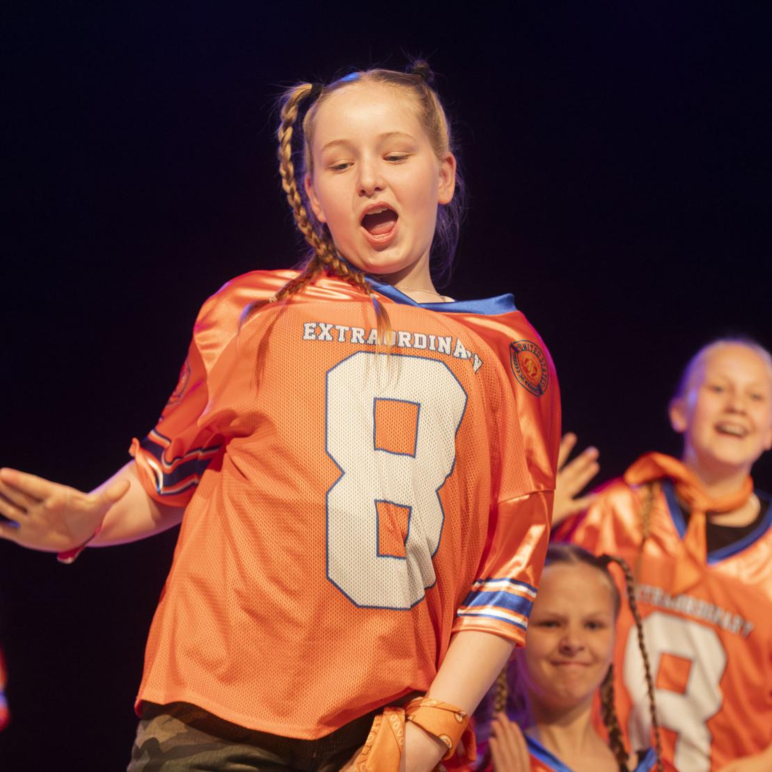 M&auml;dchen in orangefarbener Sporttrikots tanzt begeistert auf einer B&uuml;hne, umgeben von weiteren T&auml;nzern.