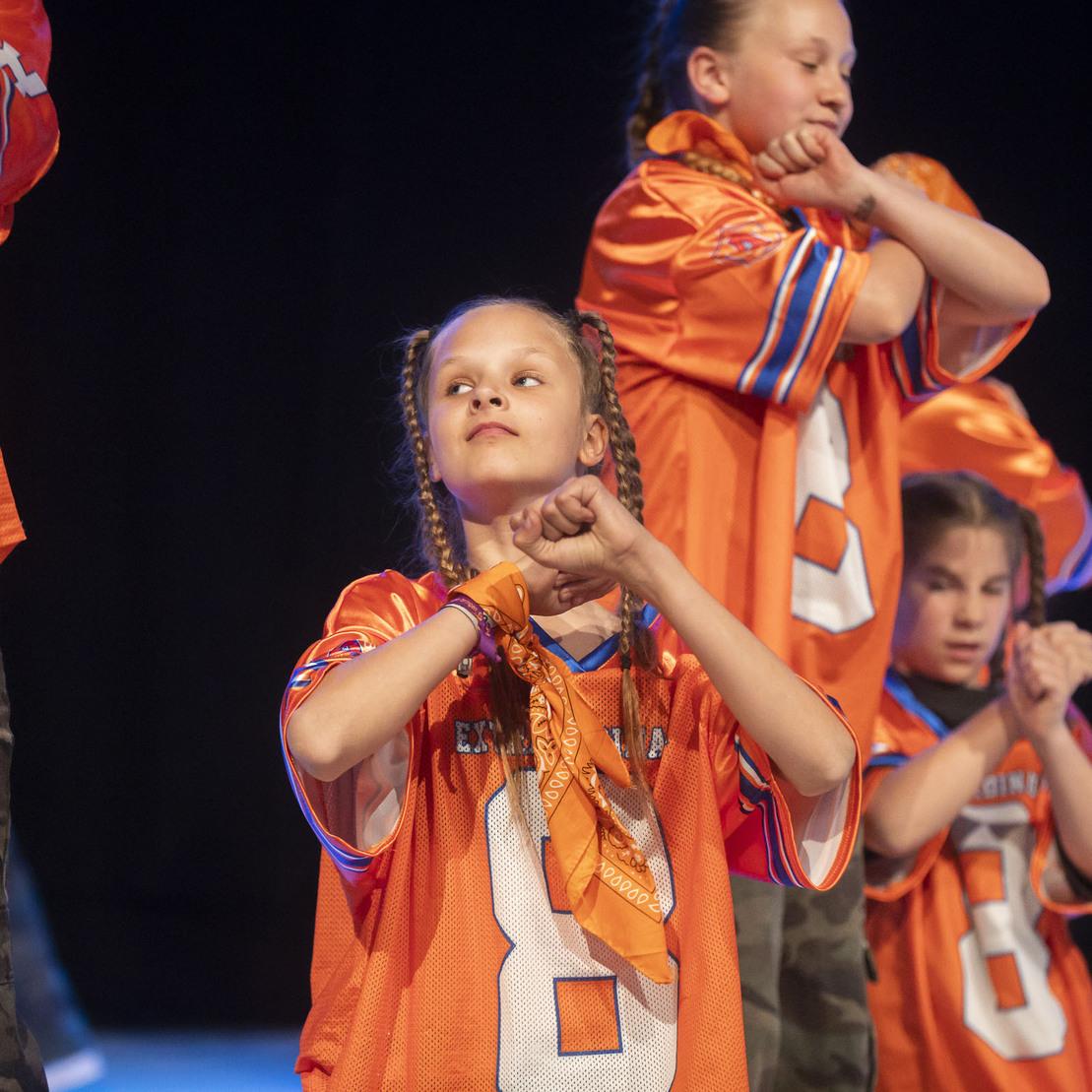 M&auml;dchen in orangefarbener Sportkleidung f&uuml;hrt eine Choreografie auf einer B&uuml;hne mit anderen T&auml;nzern aus.