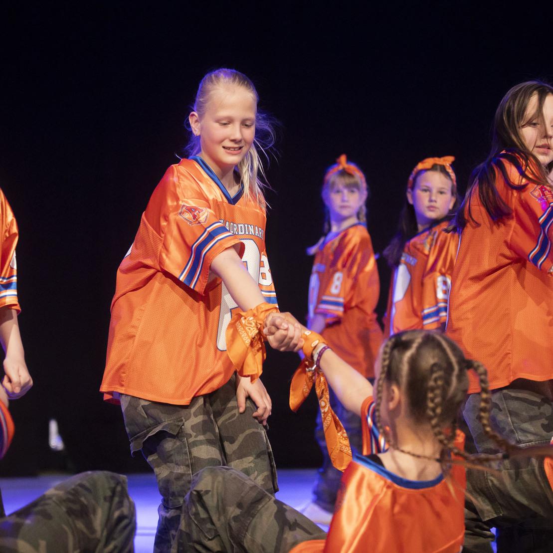 Tanzende M&auml;dchen in orangen Trikots und camouflage Hosen f&uuml;hren eine Gruppenperformance auf einer B&uuml;hne durch.